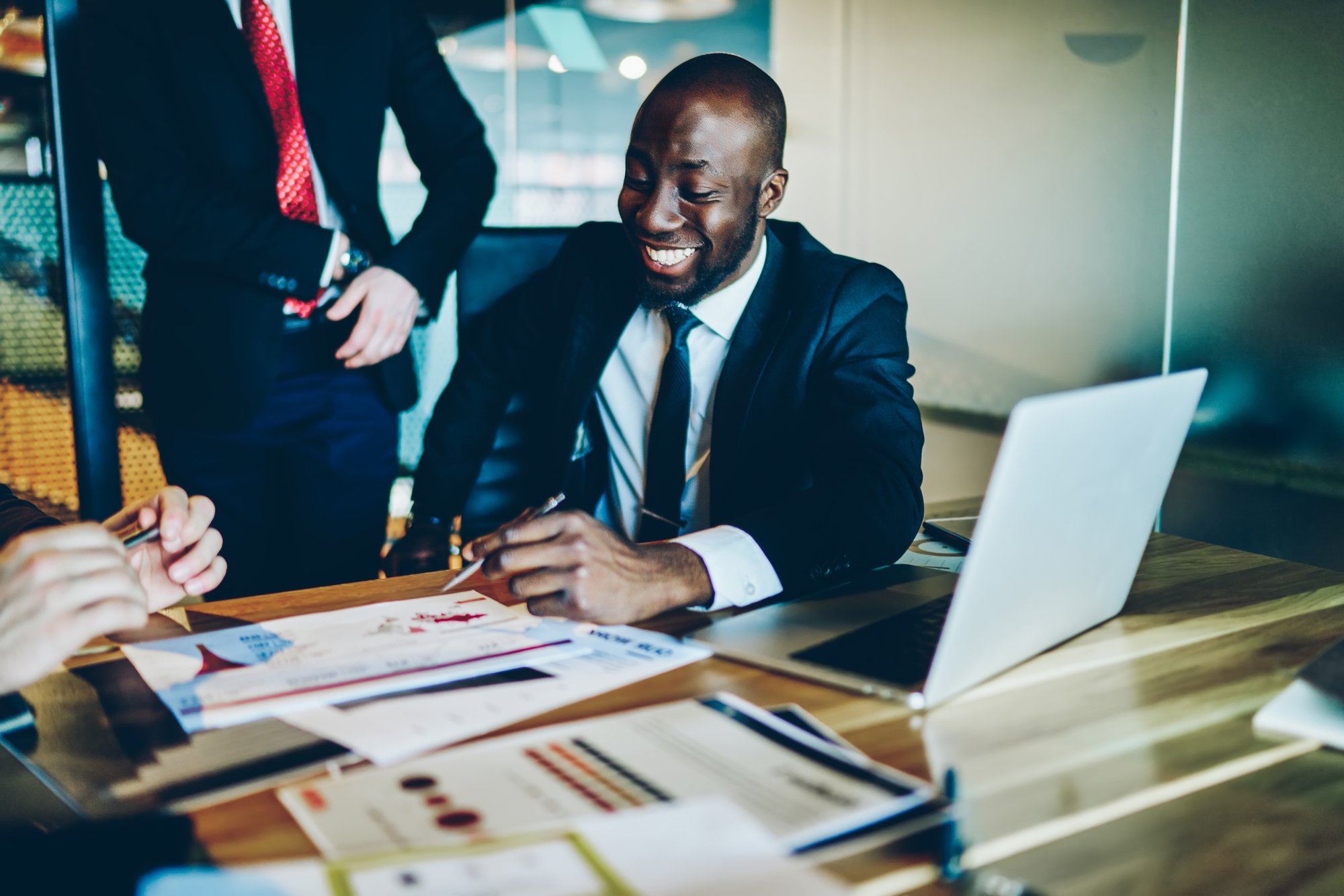 Cheerful dark skinned employee enjoying friendly collaboration with cropped colleagues discussing business strategy during paperwork, successful partners brainstorming on exchange statistics