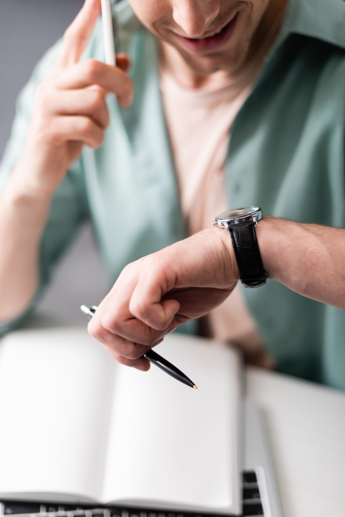 Cropped view of smiling freelancer checking time while talking on smartphone and holding pen, concept of time management