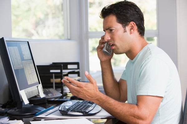 Man in home office on telephone using computer and frowning