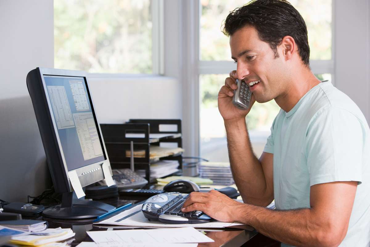 Man in home office on telephone using computer and smiling (R) (S)