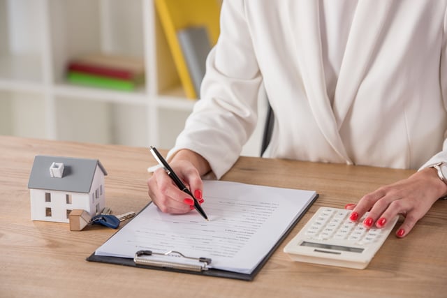 Partial view of businesswoman using calculator and writing in contract near house model and keys