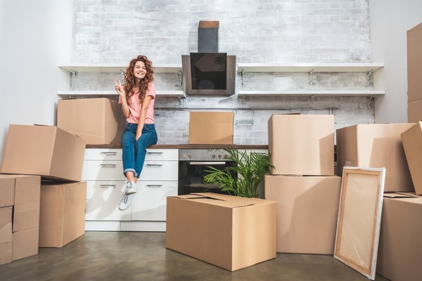 Smiling attractive woman sitting on kitchen counter between cardboard boxes and showing peace sign at new home