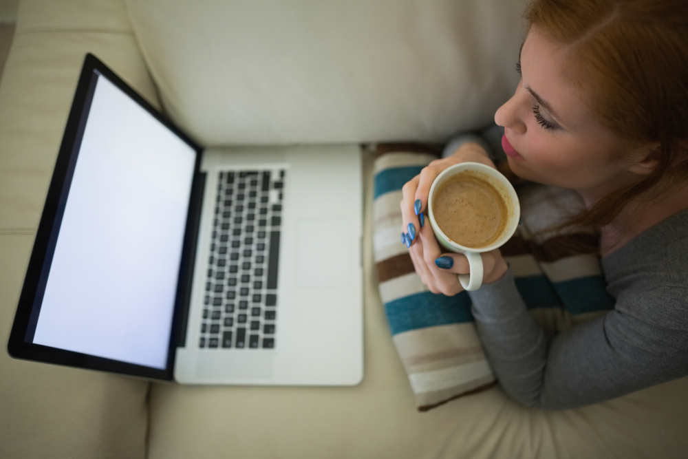 Redhead lying on the couch looking at laptop holding mug of coffee at home in the sitting room